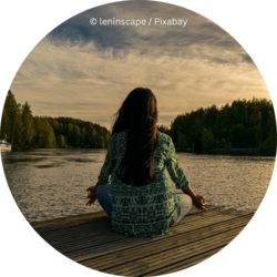 Frau sitzt im Schneidersitz auf einem Holzsteg und meditiert am Wasser bei Sonnenuntergang.