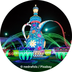 Große, bunt beleuchtete Kanne mit Blumenmustern, aus deren Ausguss rotes Licht wie Wasser fließt; umgeben von leuchtenden Tassen und farbigen Lichtbögen bei Nacht.