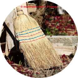 Ein handgemachter Strohbesen mit roten und blauen Zierfäden lehnt an einem Holzstapel im Freien, umgeben von verstreuten roten Beeren.