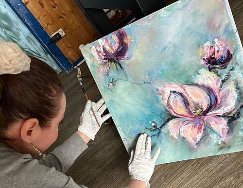 Eine Frau mit zusammengebundenen Haaren und weißem Haargummi hält ein gemaltes Blumenbild auf dem Boden fest. Sie trägt weiße Handschuhe und arbeitet an der Leinwand mit einem feinen Pinsel. Das Bild zeigt drei große, pastellfarbene Blüten auf blaugrünem Hintergrund.