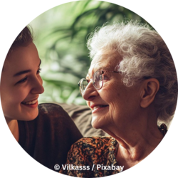 Eine ältere Frau mit Brille und grauen Haaren lächelt eine junge Frau an; beide blicken sich herzlich an und sitzen nah beieinander.