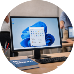 Desktop-Computer mit Windows-11-Startmenü auf dem Bildschirm; davor Tastatur und ein Buch mit dem Titel „PC Lehrbuch“, im Hintergrund ein unscharfer Schulungsraum.
