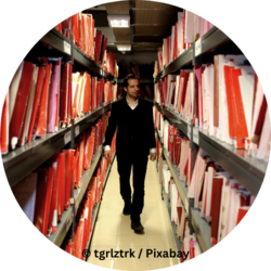 Mann geht durch einen schmalen Archivgang zwischen hohen Regalen voller roter und weißer Aktenordner.