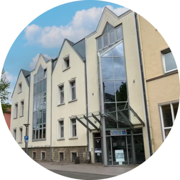 Modernisiertes, helles Gebäude mit großen Glasflächen und markantem Eingangsbereich, Schild mit „VHS“ über dem Eingang.