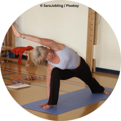 Eine Frau macht auf einer Yogamatte in einer Turnhalle eine seitliche Dehnübung mit ausgestrecktem Arm.