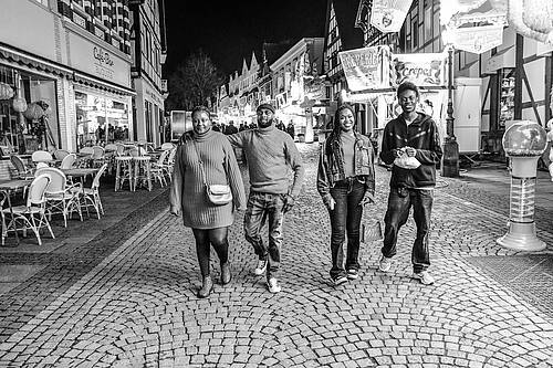 Vier junge Menschen gehen lachend nebeneinander auf einer gepflasterten Straße in einer abendlichen Altstadt, im Hintergrund leuchten Schilder und Cafés. Die Aufnahme ist in Schwarz-Weiß.