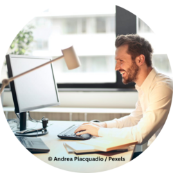 Ein lächelnder Mann mit Bart sitzt an einem Schreibtisch und arbeitet am Computer. Tageslicht fällt durch ein Fenster im Hintergrund. Auf dem Tisch liegen Bücher, eine Tastatur und eine Schreibtischlampe.