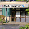 Eingang des Medienzentrums Schaumburg mit Schild über der Tür, umgeben von grünen Büschen und Bäumen.