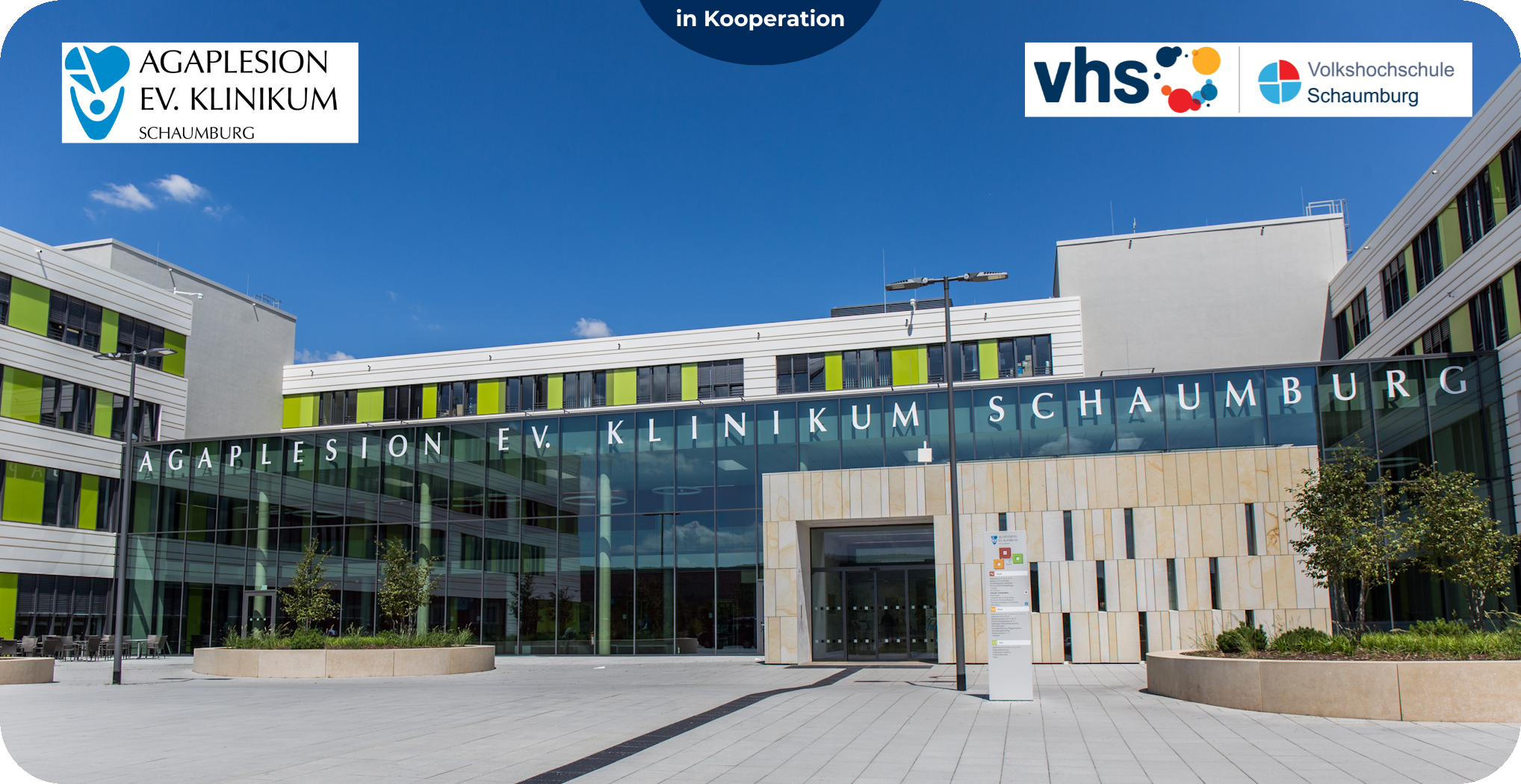 Moderne Glasfassade des AGAPLESION Evangelischen Klinikums Schaumburg bei Sonnenschein, mit Logos der Kooperationspartner AGAPLESION und VHS Schaumburg am oberen Bildrand.